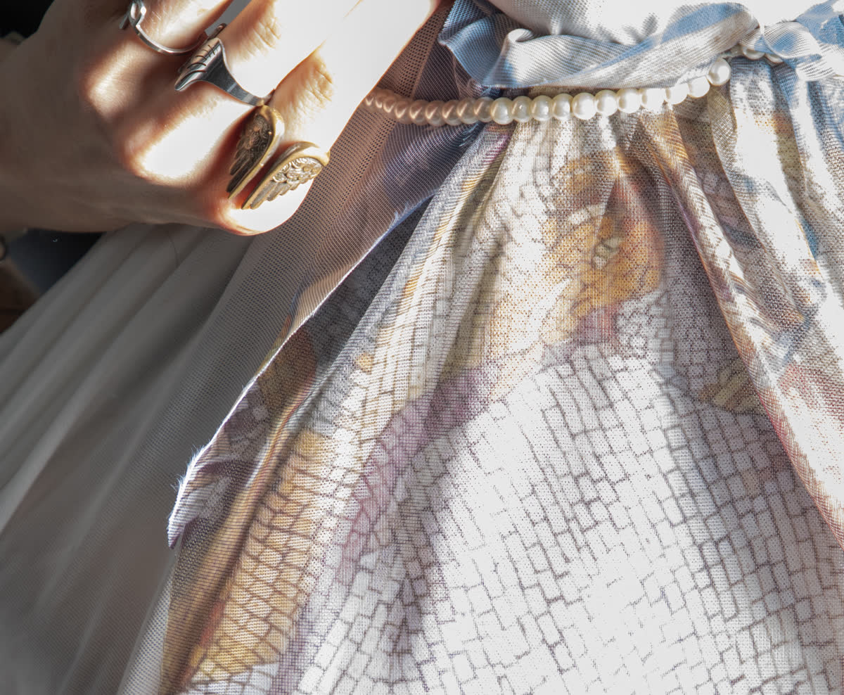 a hand ringed in silver and gold resting on sheer printed fabric, a string of pearls at the gather, pale florals under a fine grid