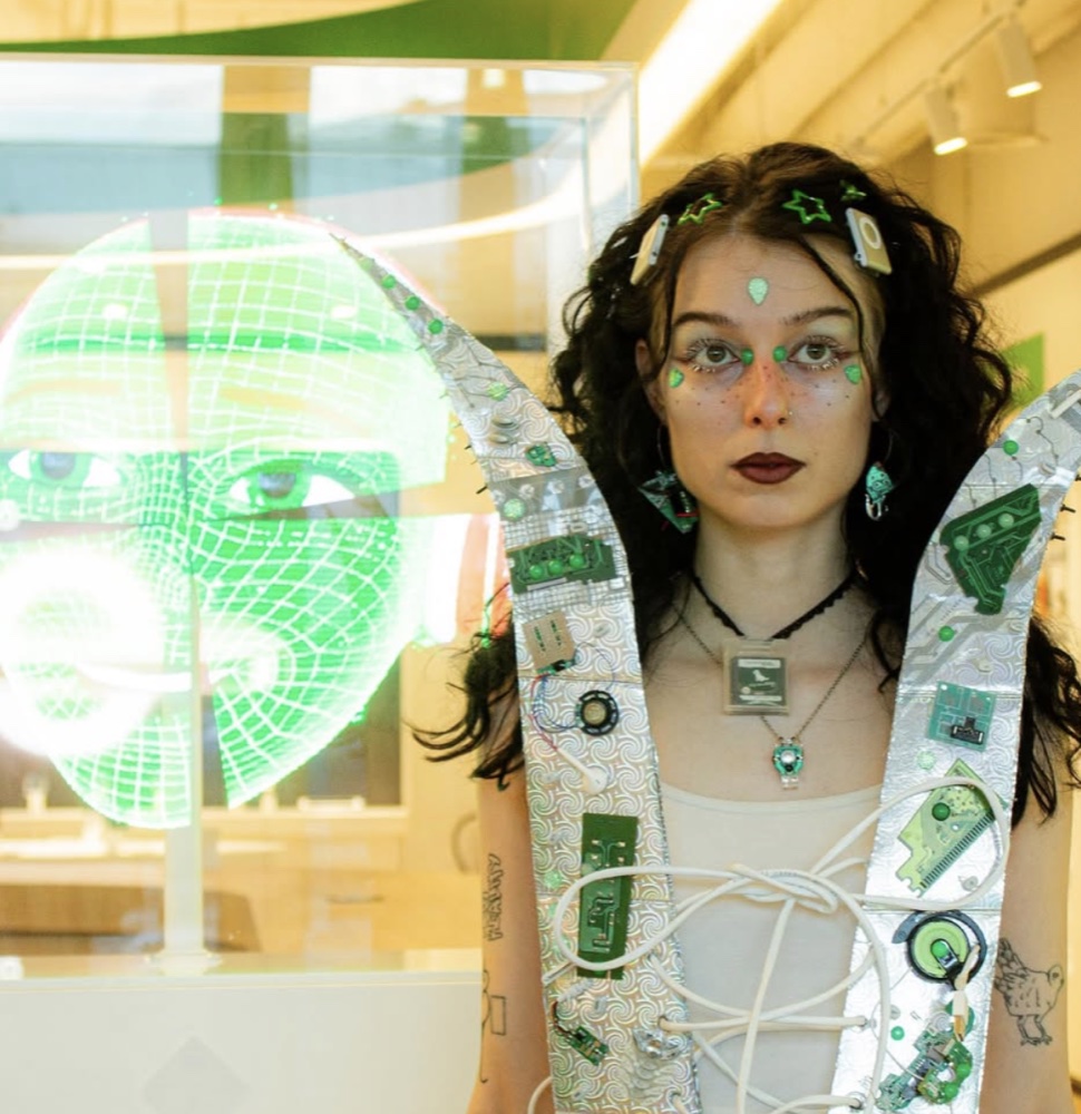 cyberbirdxx in a couture look built from circuit boards and green gems, a wireframe face projected on glass behind her