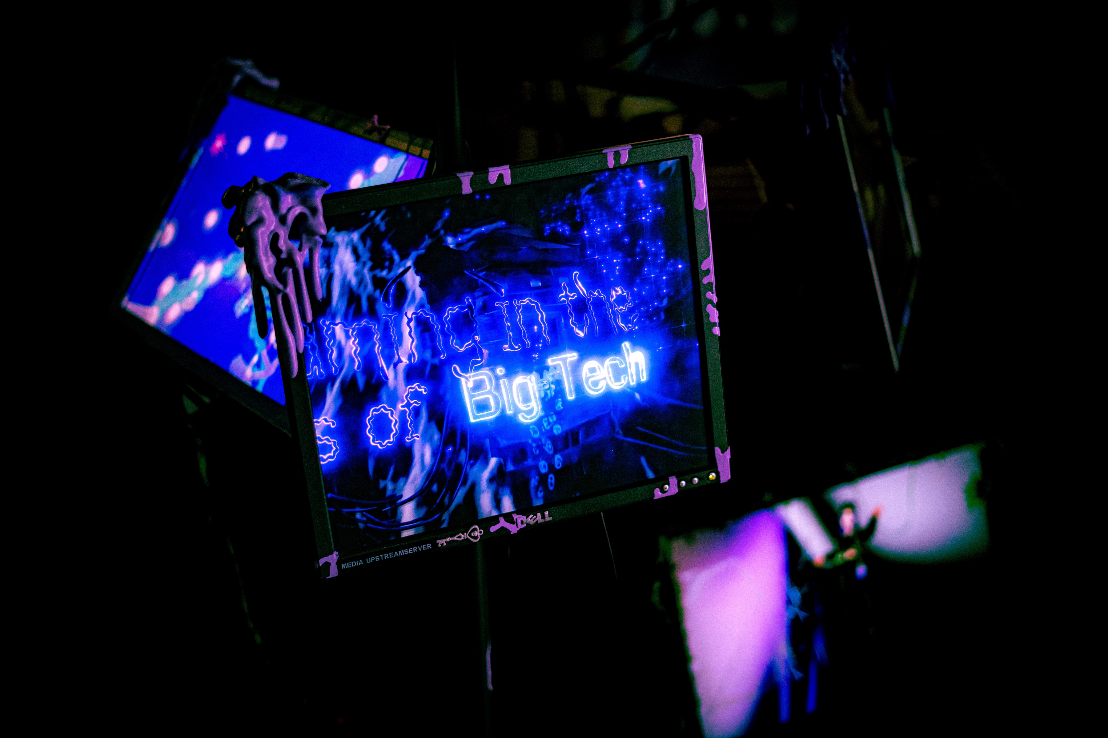 a monitor on a black pole seen at an angle, magenta slime on the frame, the screen showing the title again, 'of big tech' in bright blue lettering over blue sparks and a blurred rig, more panels glowing purple around it