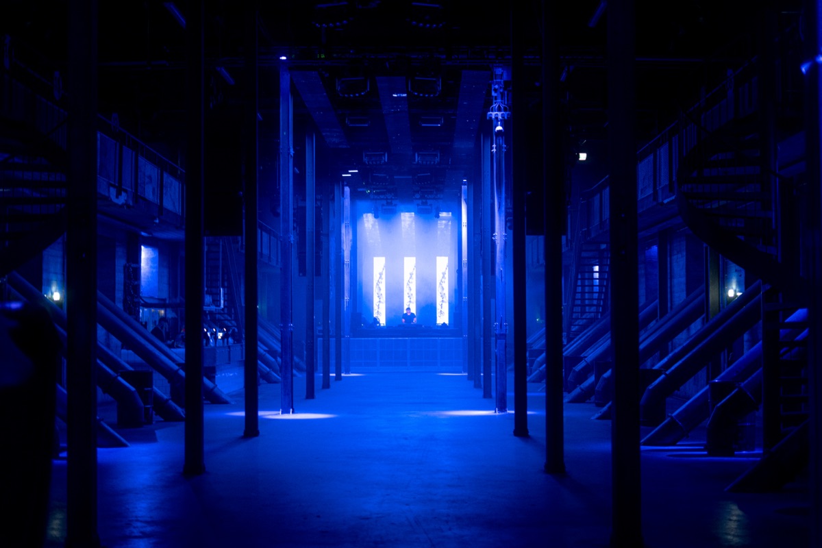 a concrete hall lit deep blue, twelve led panels around a dj at the far end