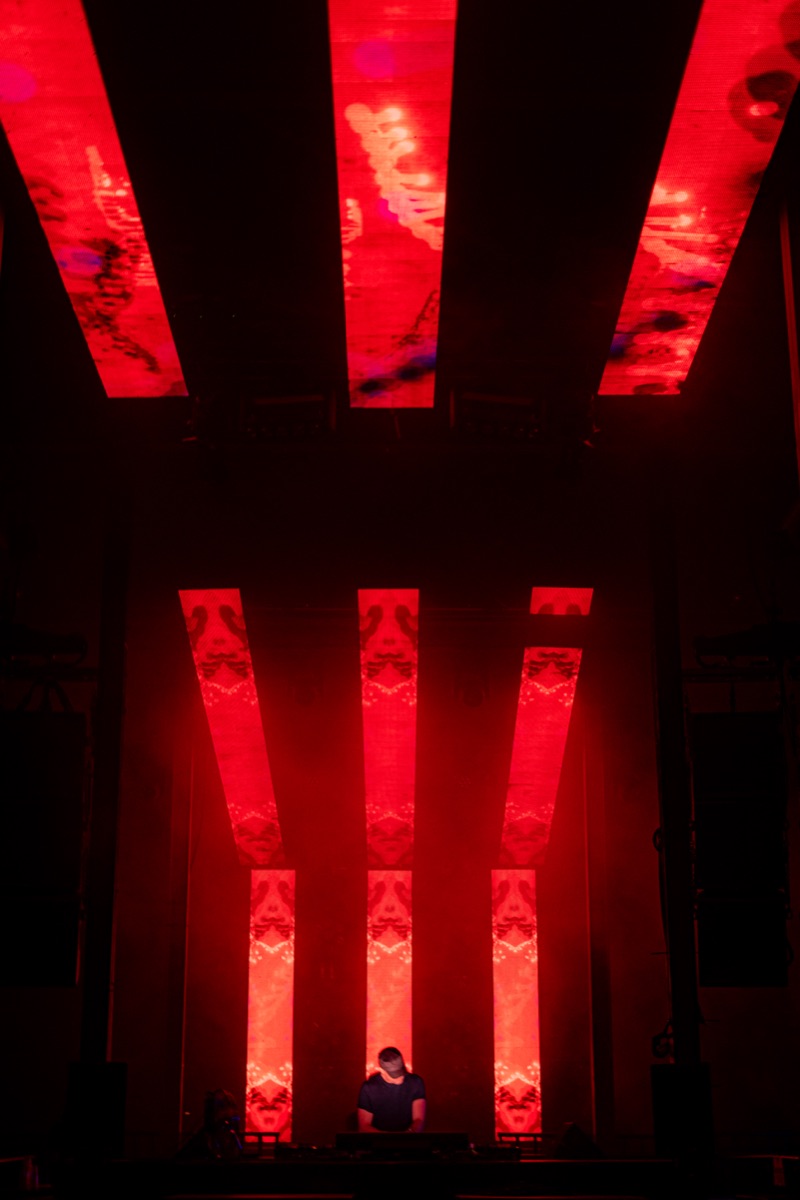 three vertical led panels glowing red, showing a mirrored symmetrical pattern like a sigil, a dj silhouetted below.