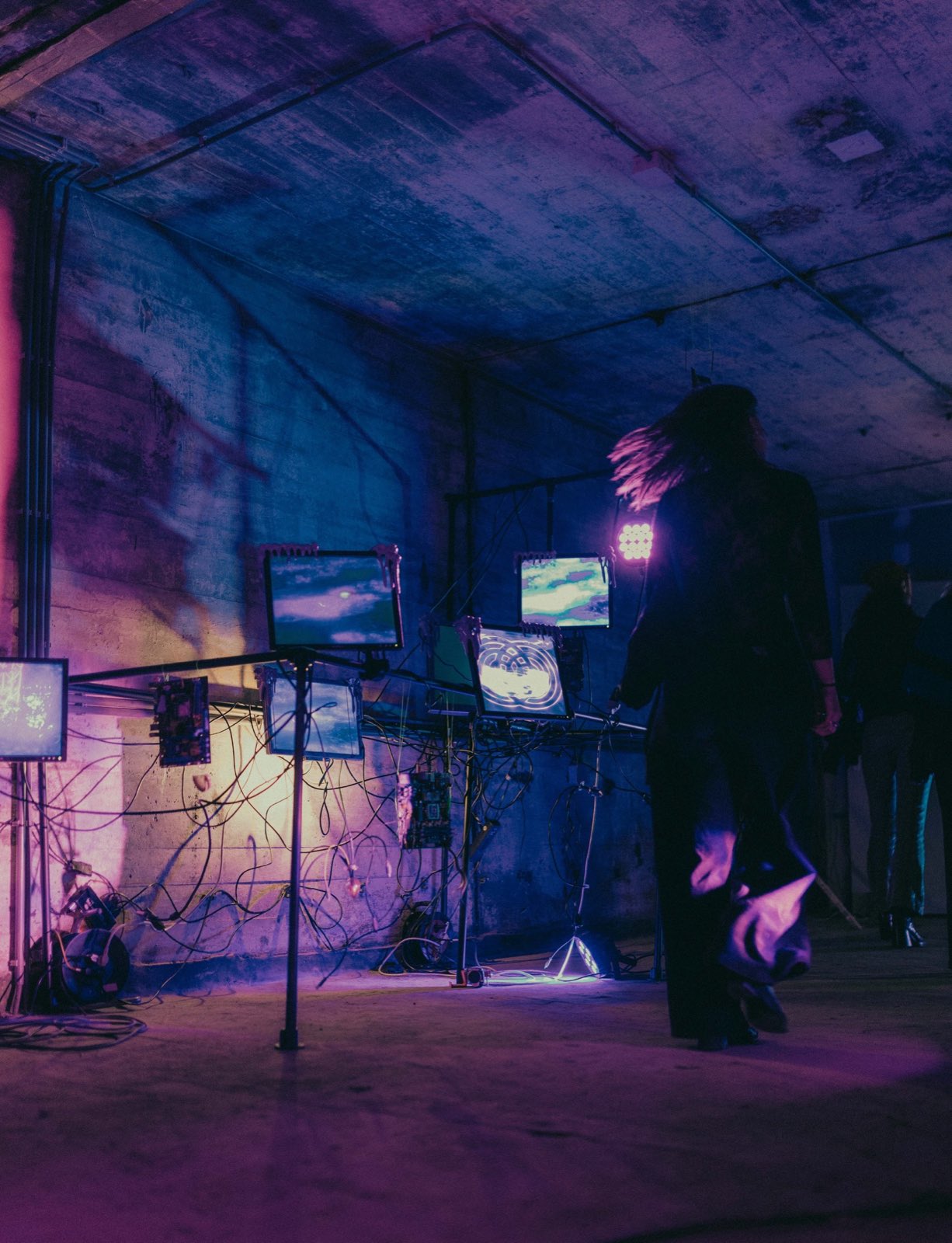 the same rig seen from further right. a person in a dark coat walks past in the foreground, hair caught in motion blur. the monitors show blurred water surfaces and one shows a glowing knotwork spiral. other figures stand in shadow at the right.