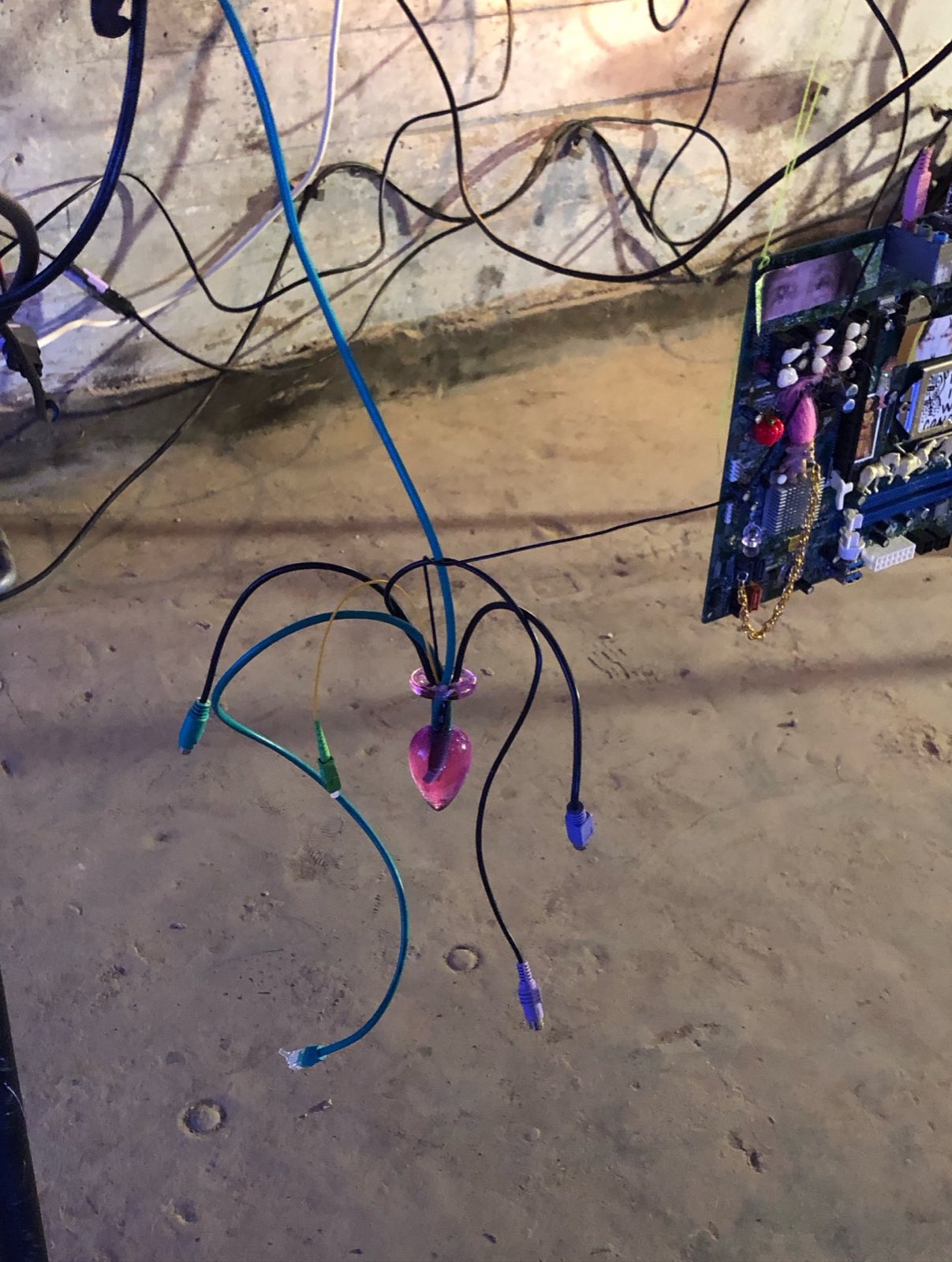a translucent pink heart shaped object hangs at the end of a bundle of cables just above a dusty concrete floor. a blue ethernet cable passes through the flat disc at its top. loose unplugged connectors lie on the floor nearby. at right, a circuit board hung on the wall carries a gold chain, white beads, a red strawberry charm and small printed photographs.