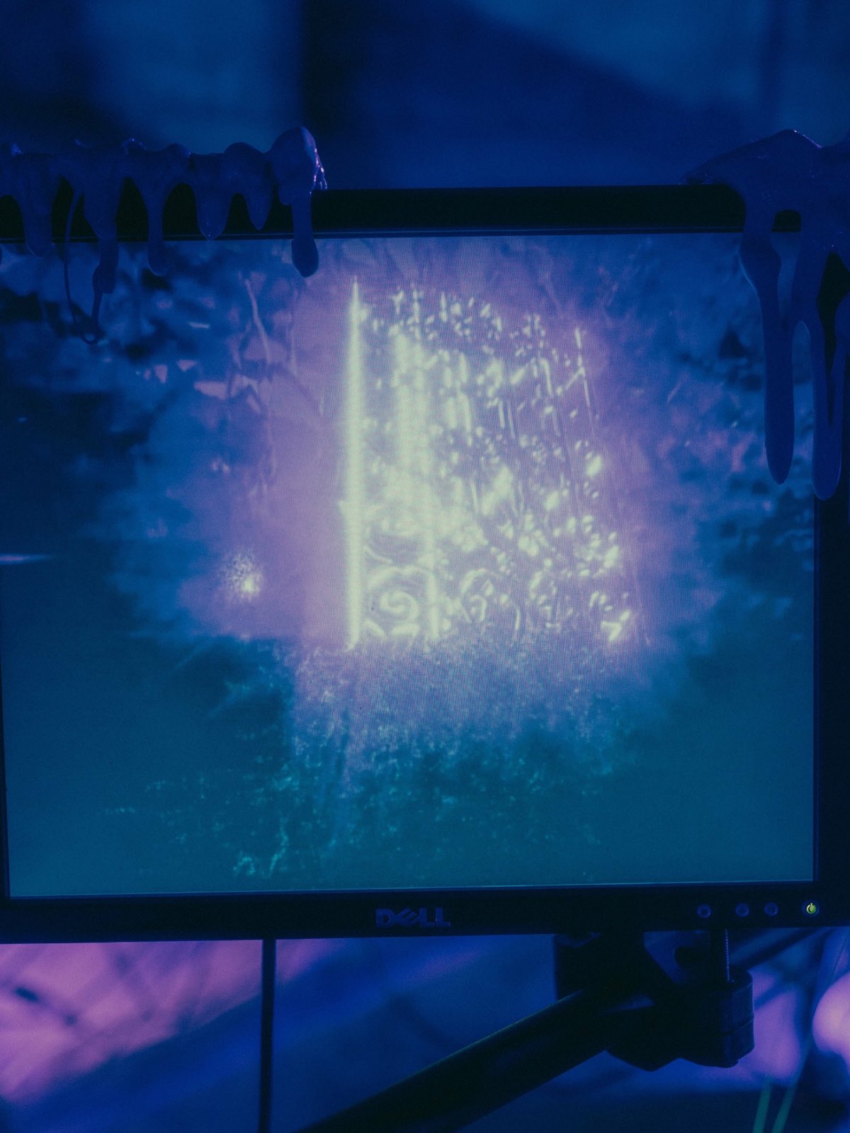 a dell monitor filling the frame. pale cyberslime drips hang over its top bezel. the screen shows a bright vertical column of light rising through dark water, with pale specks and stems around it, pink above and green below.