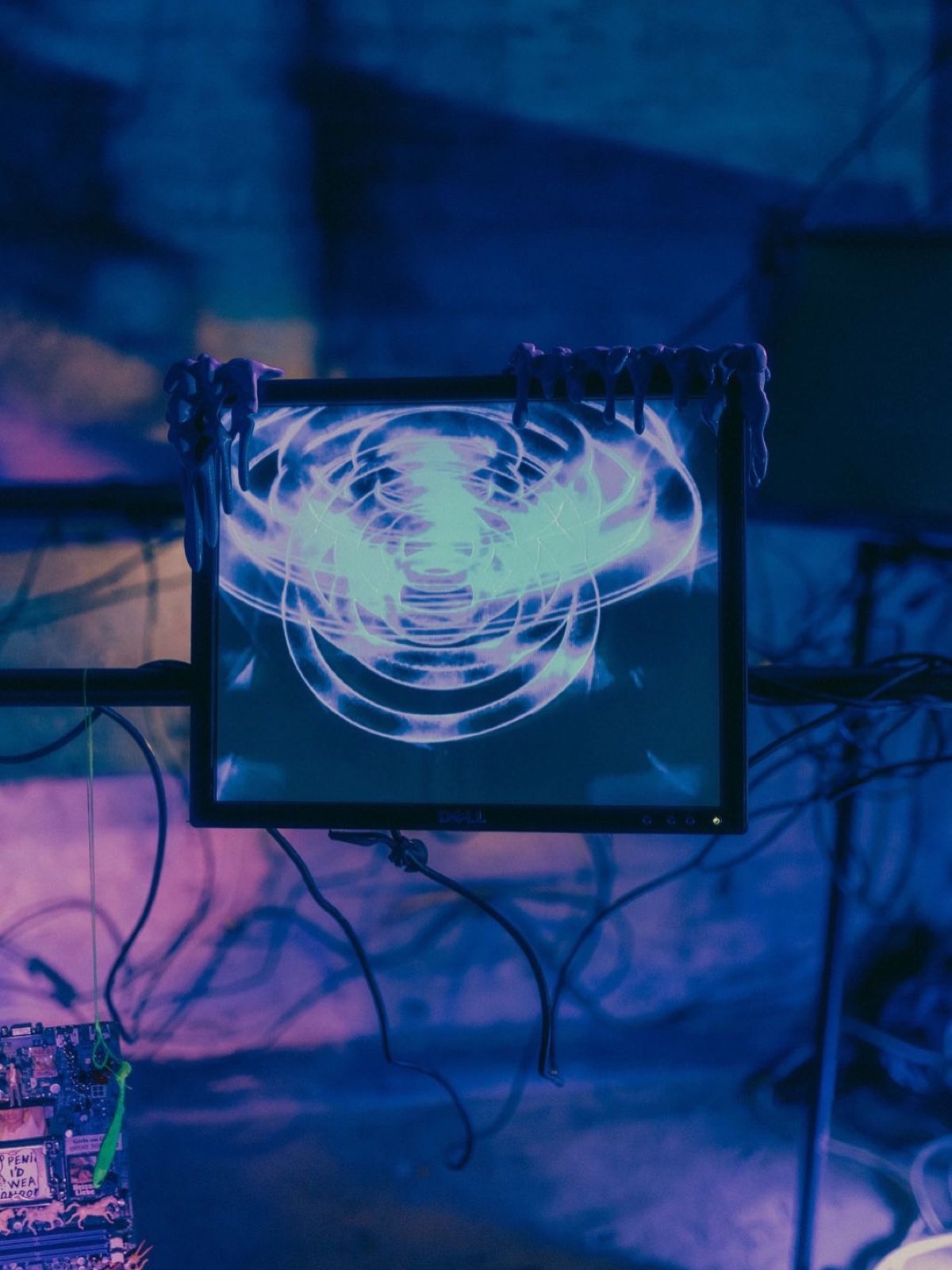 a dell monitor mounted on a black pole against a violet wall. the screen shows large concentric rings in glowing pale green on dark blue, a water ripple pattern doubled and torn open. a decorated circuit board with a green plastic charm on lime string hangs at the lower left.