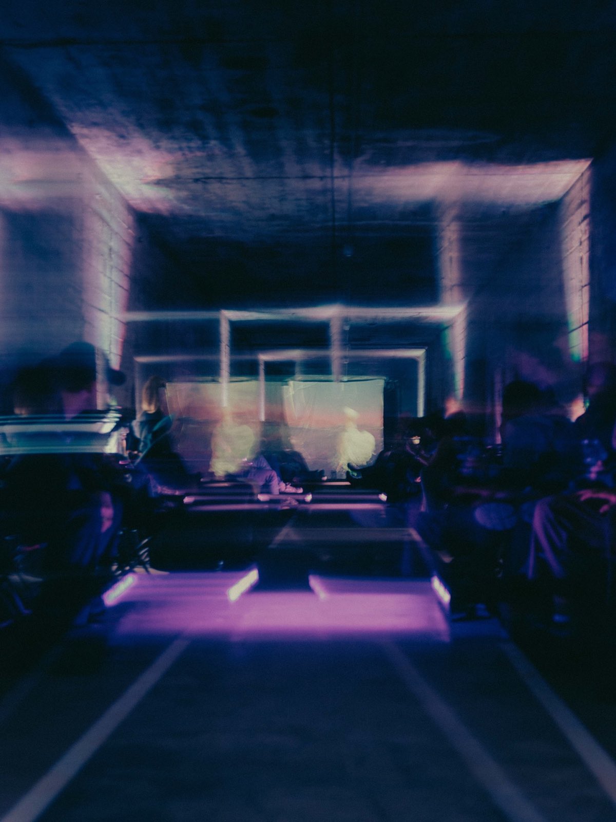 the hall seen down its length in low light. a raised runway with lit edges runs into the room, with a pool of magenta light across it. an audience sits either side. pale yellow translucent screens stand at the far end.