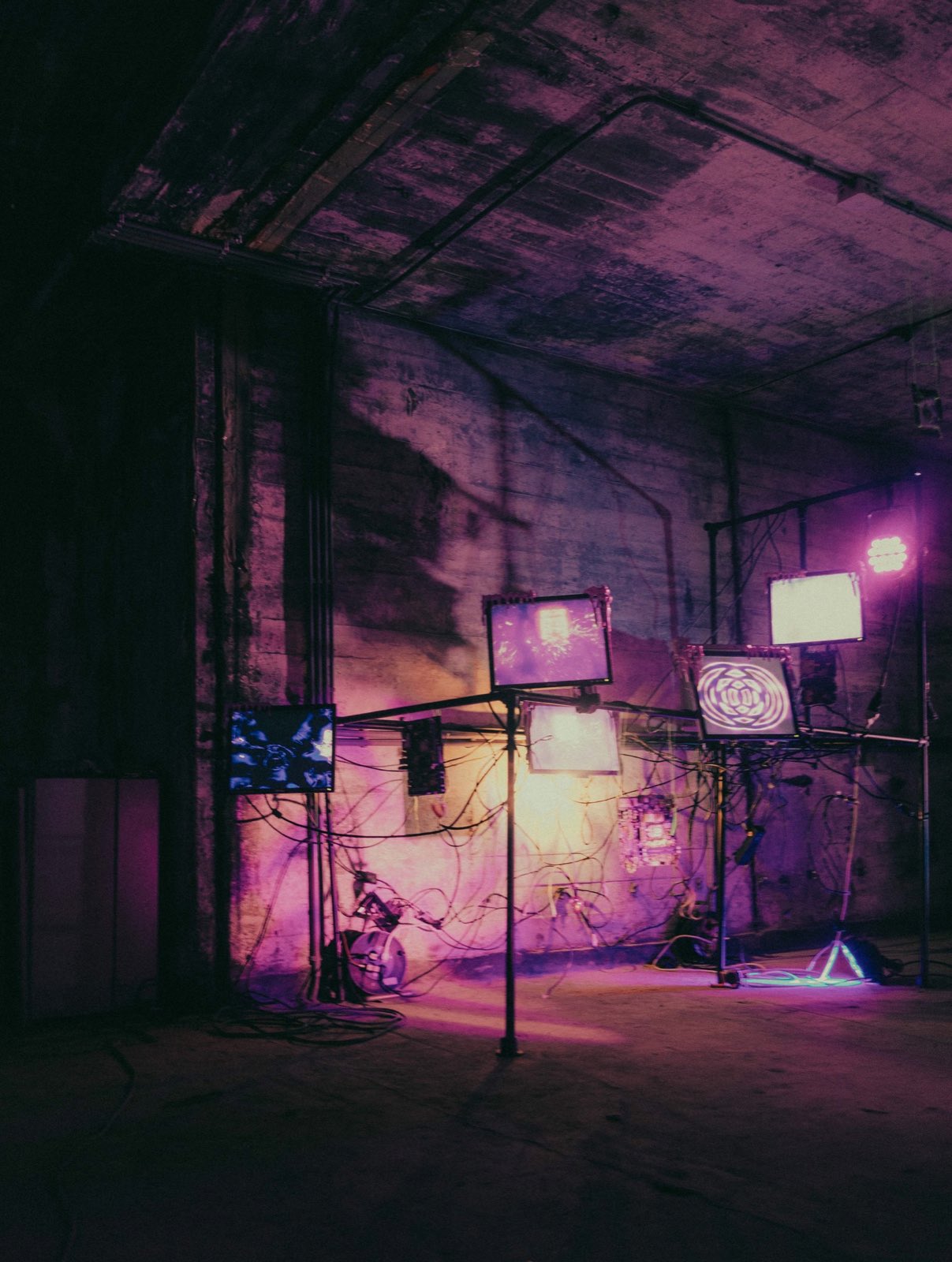 the rig against the concrete wall, the monitors lit. one shows white marbled reflections, another a pale wash, another a bright white panel, another a concentric ripple rosette. a magenta lamp on a stand at right and a small white lamp on the floor light the wall. a warm amber wash falls behind the cables.