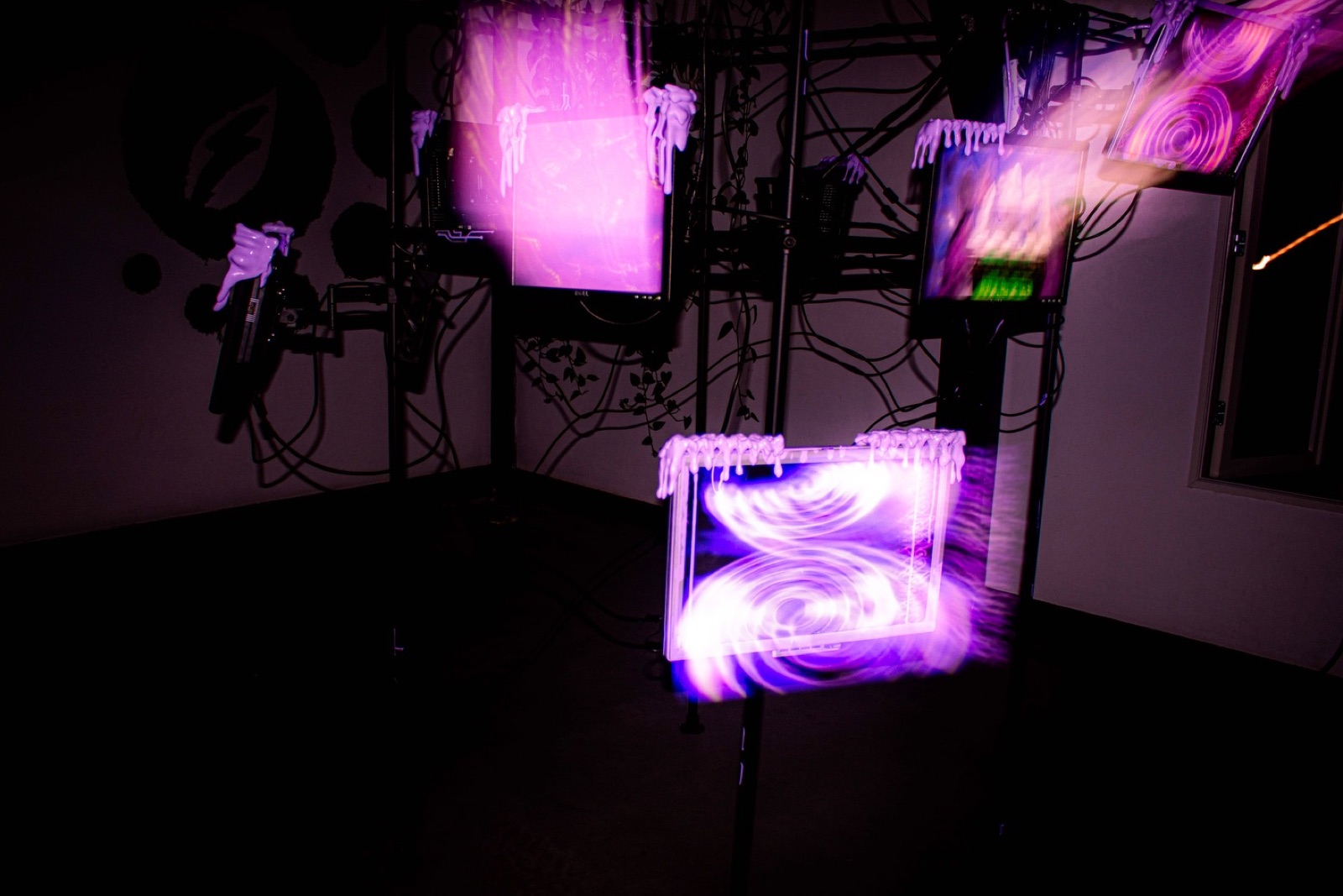 a wider view of the rig in a dark room, several monitors with cyberslime dripping over their bezels showing pink and violet visuals, a moulded white hand mounted on the wall at left holding a spray gun.