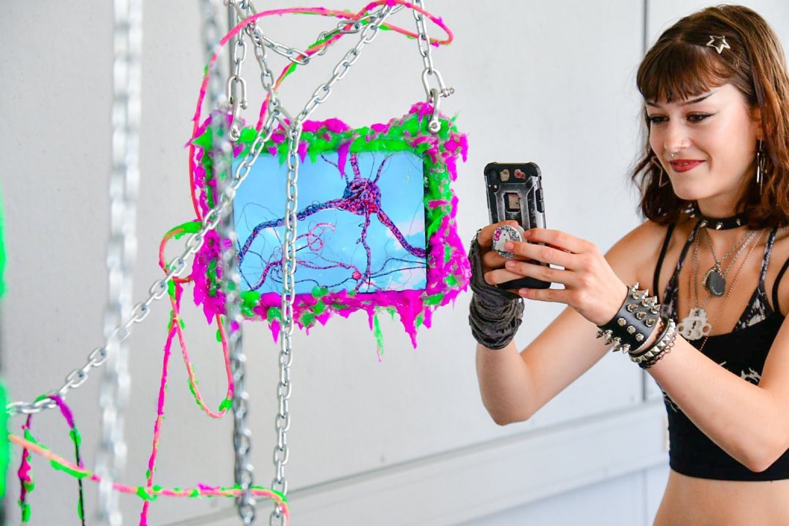 a visitor photographs a small screen on her phone, the screen bordered in dripping magenta and green cyberslime and hung on steel chain, showing a tangled blue and pink circuit-like drawing.